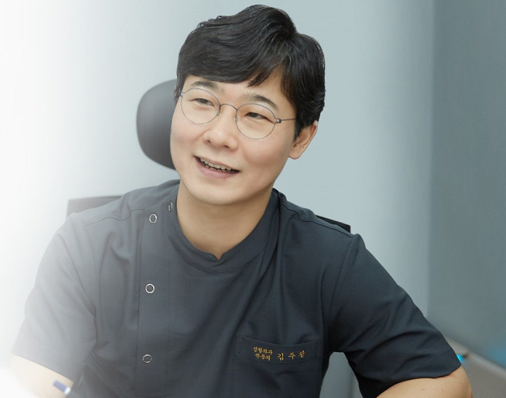진료실 책상에서 웃으며 상담하는 대표원장 김주찬(가운에 성형외과 전문의 김주찬 수)
